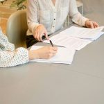 Negotiation - woman signing on white printer paper beside woman about to touch the documents